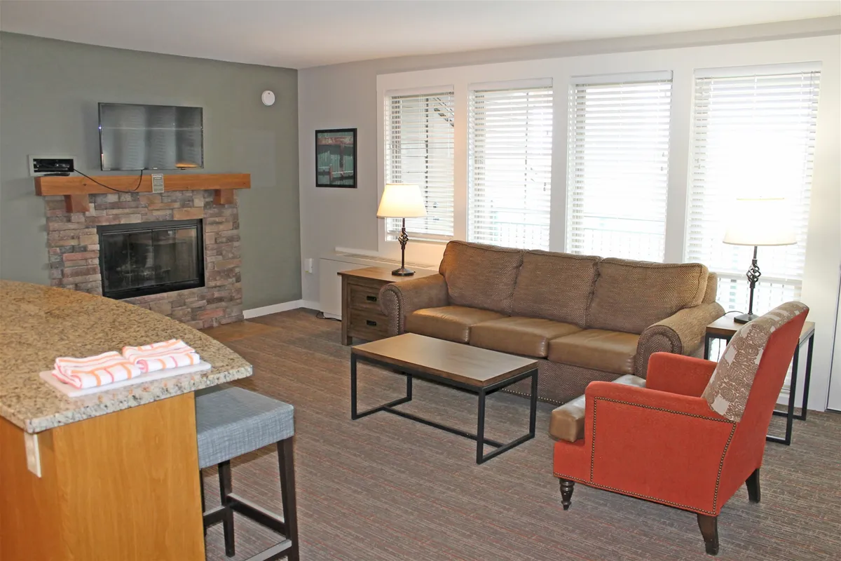 The image shows a warm, inviting living area featuring a glowing fireplace, a comfortable sofa, a plush armchair, and a coffee table set for relaxation or shared meals. Sunlight softly streams through the windows, enhancing the cozy atmosphere and highlighting the homey, resort-style furnishings.