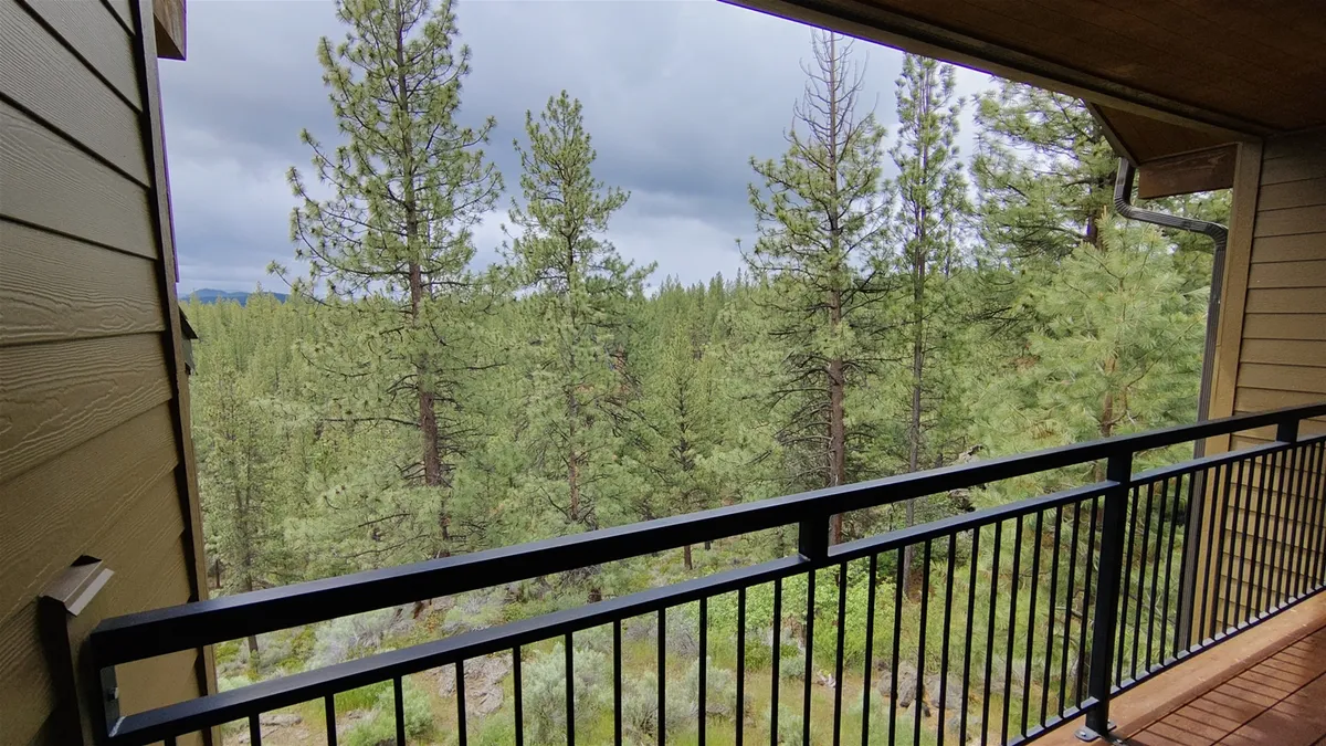 This image provides a sweeping view from the balcony, featuring a sturdy railing framing tall pine trees and distant mountain peaks. The scene conveys the natural beauty that surrounds the property and a sense of tranquil seclusion, with the vantage point suggesting a private, elevated perch perfect for enjoying the outdoors.