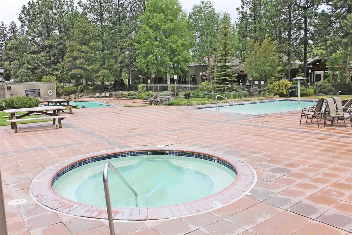 This exterior image captures the resort's outdoor amenities, including a sparkling swimming pool, inviting hot tub, picnic tables, and lush green trees surrounding the area. The scene suggests a vibrant, active atmosphere where families can enjoy swimming, relaxing together, and socializing in the beautiful natural setting.