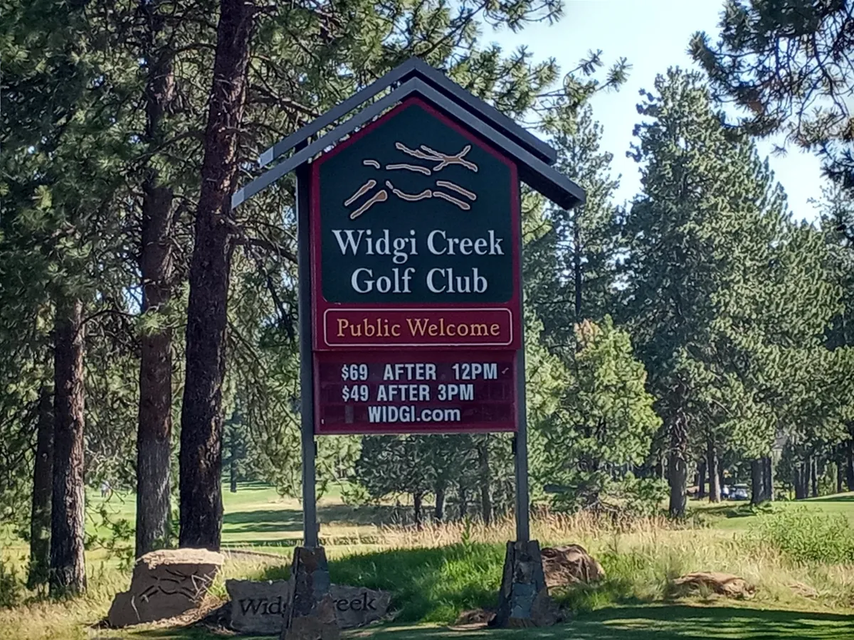 The image highlights a golf club sign nestled among natural surroundings, with some price information visible. The area is open and inviting with greenery and the promise of scenic golf courses nearby.