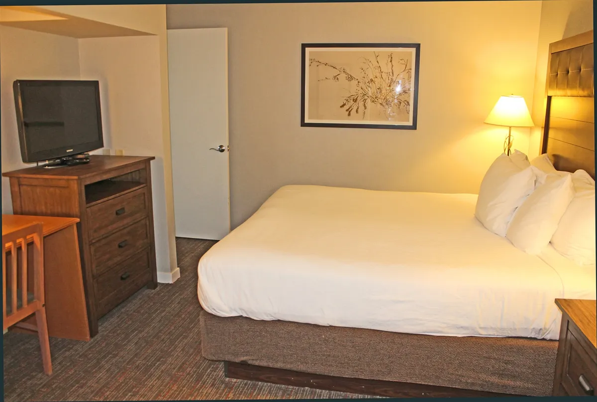A serene king bedroom featuring a plush bed, soft lamp lighting, wall-mounted TV, and alpine-inspired artwork. The ambiance is tranquil and relaxed, suggesting a perfect retreat for rest or quiet reading after a day of adventure.