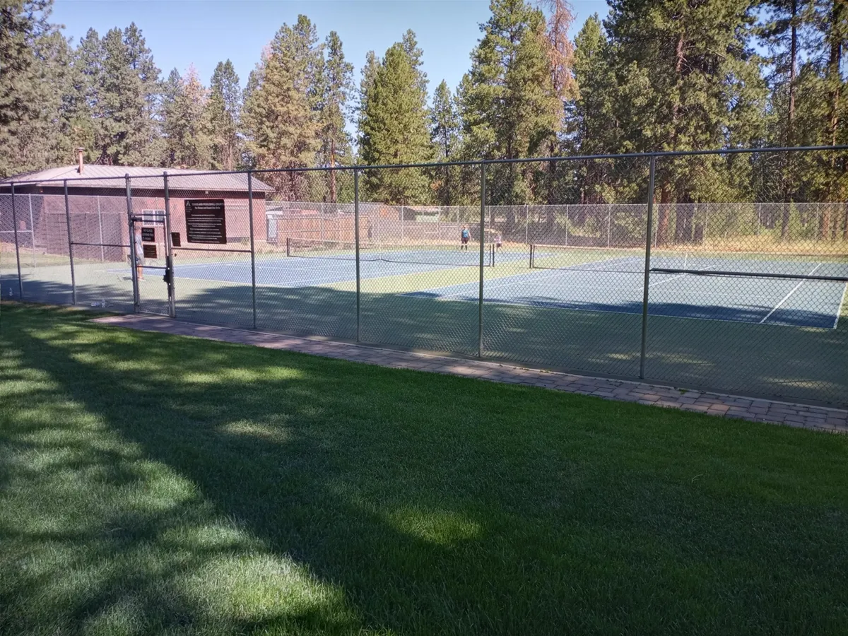 Pristine tennis courts bordered by lush green grass and tall trees, with a clubhouse in the background. The courts are well-maintained, ready for friendly matches or organized tournaments, and the surrounding environment is active and communal.