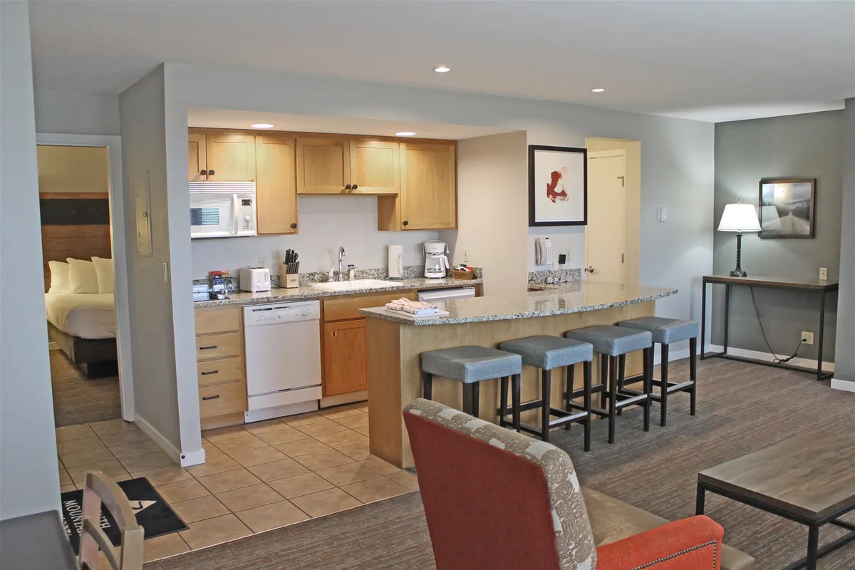 An inviting open-plan kitchen and living space featuring a kitchen island with bar stools, framed artwork on the walls, and a cozy living area for relaxing or socializing. The kitchen area suggests easy meal prep and a hub for gathering, while the connected living area hints at a comfortable, communal environment.